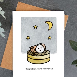 May include: A greeting card featuring a cartoon dumpling in a steamer basket under a night sky with a crescent moon and stars. The text "Congrats on your lil' dumpling" is printed below the illustration. The card is on a brown envelope.