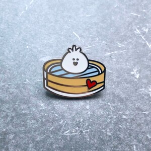 May include: A white and blue enamel pin of a cartoon dumpling with a smiling face, sitting in a yellow and brown bamboo steamer with a red heart on the side.