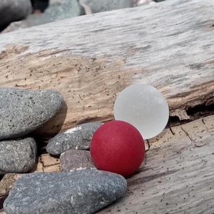 May include: A collection of smooth, gray stones and two glass spheres, one red and one white, rest on a weathered piece of driftwood. The scene is lit by natural sunlight, creating a rustic and natural aesthetic.