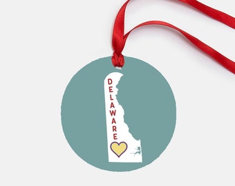 Puede incluir: Adorno navideño redondo, de color verde azulado, con una cinta roja para colgar. El adorno presenta un contorno blanco del estado de Delaware, con la palabra "DELAWARE" en rojo y un corazón amarillo.