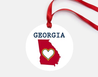 Puede incluir: Adorno redondo blanco con un contorno rojo del estado de Georgia y un corazón blanco con un contorno amarillo en su interior. El texto "GEORGIA" está impreso en azul en la parte superior del adorno.