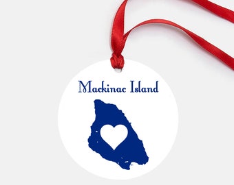 May include: White round ornament with a blue outline of Mackinac Island and a white heart in the center. The text "Mackinac Island" is written in blue cursive above the island.