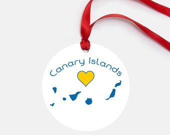 May include: White round ornament with a blue outline and the text "Canary Islands" in blue. A yellow heart is in the center of the ornament, and a blue outline of the Canary Islands is below the heart.