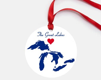 May include: A white round ornament with a blue outline of the Great Lakes and a red heart in the center. The text "The Great Lakes" is written above the heart. The ornament has a red ribbon attached.