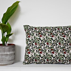 May include: A white pillow with a green and red floral pattern. The pillow is on a white shelf with a potted plant in the background.