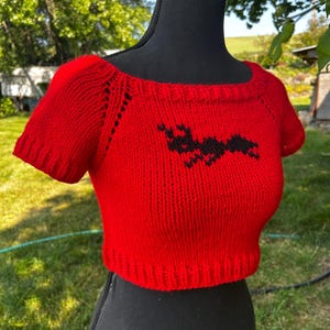 May include: A red, short-sleeved, knitted crop top with a black bat design on the front. The top has a scoop neckline and is displayed on a black mannequin. The background shows green grass and trees.
