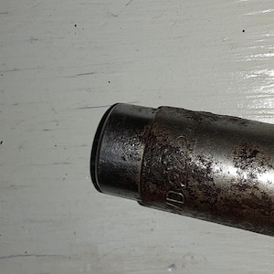 May include: Close-up of a weathered, cylindrical metal object with a dark, possibly rusted, finish. The object has a slightly flared end and appears to be a component of a larger mechanism or tool. The surface shows signs of age and use.