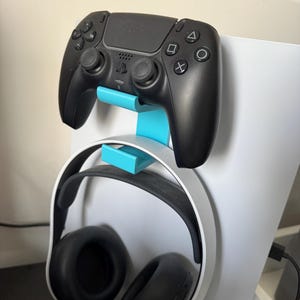 Könnte beinhalten: Ein schwarzer PlayStation 5-Controller ist auf einem hellblauen Ständer montiert, über einem weißen Headset mit schwarzen Ohrmuscheln. Das Setup befindet sich neben einer weißen PlayStation 5-Konsole.