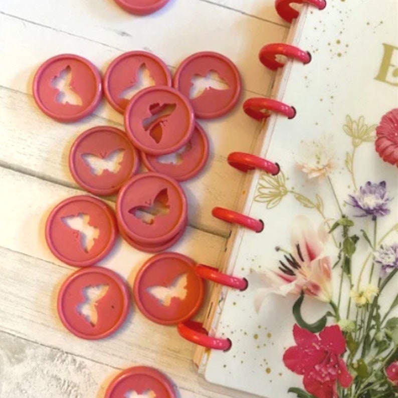 K&ouml;nnte beinhalten: Nahaufnahme einer Reihe von rosafarbenen Butterfly-Binderclips. Die Clips sind in einem Kreis auf einer wei&szlig;en Holzoberfl&auml;che angeordnet. Im Hintergrund ist ein rosa-wei&szlig;er Planer zu sehen.