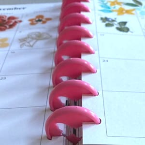 K&ouml;nnte beinhalten: Nahaufnahme eines rosafarbenen Kunststoffbindungssystems f&uuml;r einen Planer oder ein Notizbuch. Das Bindungssystem besteht aus mehreren Ringen, die miteinander verbunden sind. Die Ringe sind so konzipiert, dass sie die Seiten zusammenhalten.