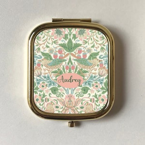 Könnte beinhalten: Ein goldener Kompakt-Spiegel mit einem Blumenmuster und dem Namen "Audrey" in einem rosa Oval. Der Spiegel hat einen weißen Hintergrund mit grünen, rosa und blauen Blumendesigns.