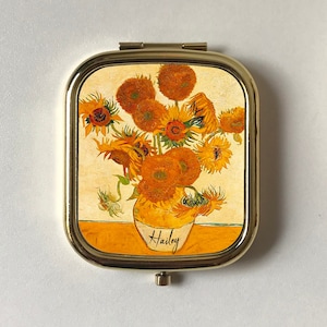 May include: A gold-colored compact mirror with a hinged lid. The lid features a painting of a vase of sunflowers, with the word "Hailey" written on the vase.