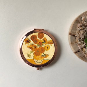 Peut inclure: Un miroir compact rond de couleur or avec un motif de tournesols dans un vase. Le miroir porte le nom "Sarah" inscrit sur le vase.