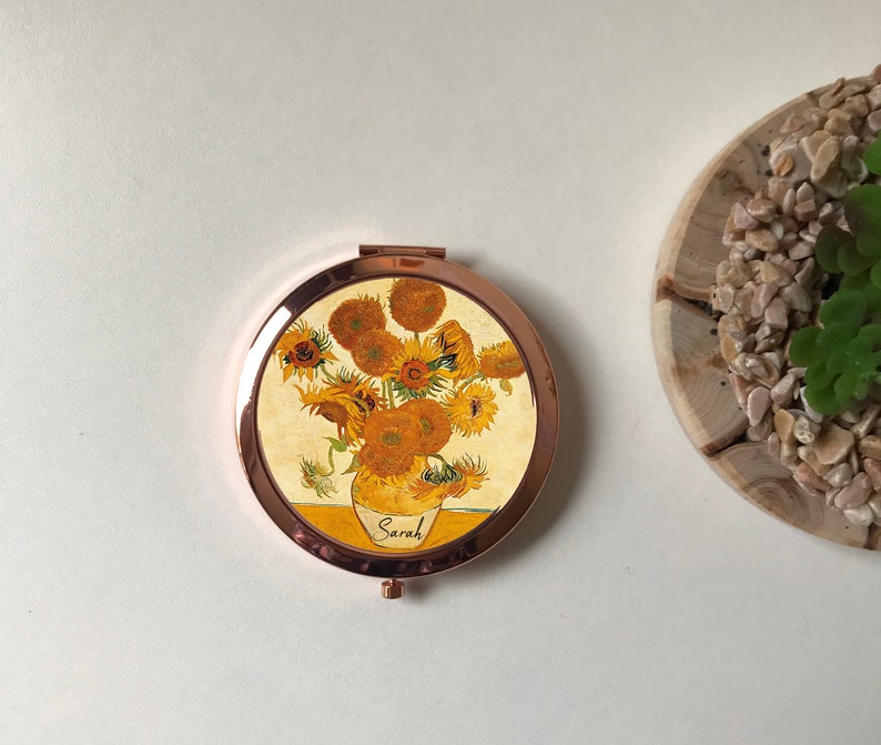 Peut inclure: Un miroir compact rond de couleur or avec un motif de tournesols dans un vase. Le miroir porte le nom "Sarah" inscrit sur le vase.