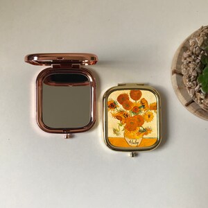 Peut inclure: Deux miroirs de poche, l'un avec une finition or rose et un miroir clair, l'autre avec une finition dor&eacute;e et une image imprim&eacute;e de tournesols dans un vase. L'image de tournesol est intitul&eacute;e "Van Gogh".