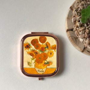 Peut inclure: Un miroir de poche avec un cadre en or rose et un motif de tournesols dans un vase. Le miroir porte le nom "Hailey" en bas.