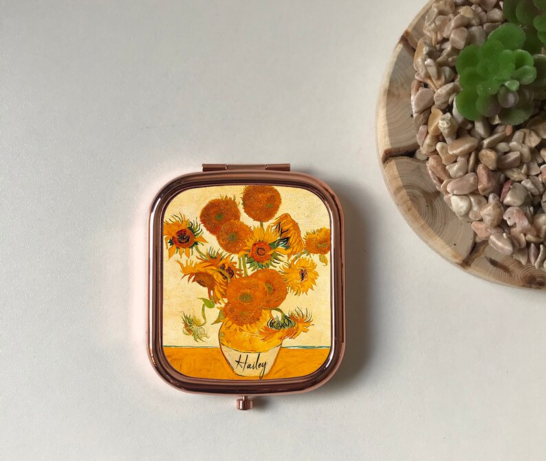 Peut inclure: Un miroir de poche avec un cadre en or rose et un motif de tournesols dans un vase. Le miroir porte le nom "Hailey" en bas.