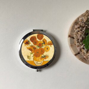 Peut inclure: Un miroir de poche rond en argent avec un motif repr&eacute;sentant une peinture de tournesols dans un vase. Le miroir porte le nom "Sarah" imprim&eacute; en bas.