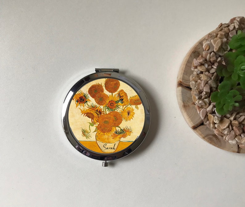 Peut inclure: Un miroir de poche rond en argent avec un motif repr&eacute;sentant une peinture de tournesols dans un vase. Le miroir porte le nom "Sarah" imprim&eacute; en bas.