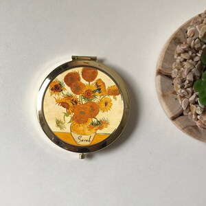 Peut inclure: Un miroir compact de forme ronde, de couleur dor&eacute;e. Le miroir pr&eacute;sente une image imprim&eacute;e d'un vase de tournesols, avec le mot "Sarah" &eacute;crit sur le vase.