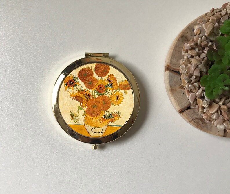 Peut inclure: Un miroir compact de forme ronde, de couleur dor&eacute;e. Le miroir pr&eacute;sente une image imprim&eacute;e d'un vase de tournesols, avec le mot "Sarah" &eacute;crit sur le vase.