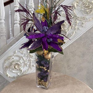 May include: A purple flower arrangement with gold accents in a clear glass vase. The vase is filled with gold beads and purple berries.