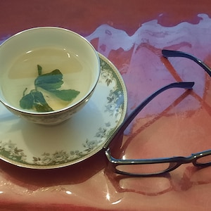 Pode incluir: Uma chávena de chá branca com um desenho floral está sobre um pires a condizer. A chávena está cheia de um líquido amarelo claro e um ramo de hortelã verde. Um par de óculos de armação preta está sobre uma superfície vermelha.