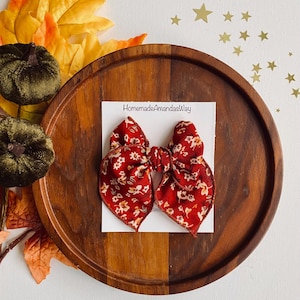 May include: A red and white floral patterned hair bow with a brown elastic band. The bow is on a white card with the text "HomemadeAmandasWay".