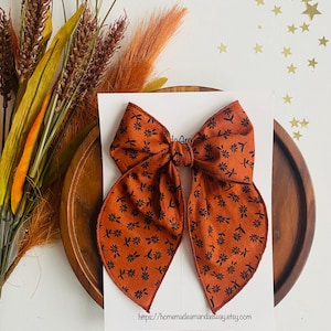 May include: A large, brown bow with a black floral pattern. The bow is made of fabric and has a slightly frayed edge. The bow is sitting on a white card with the text "https://homemadeamandasway.etsy.com" printed on it.