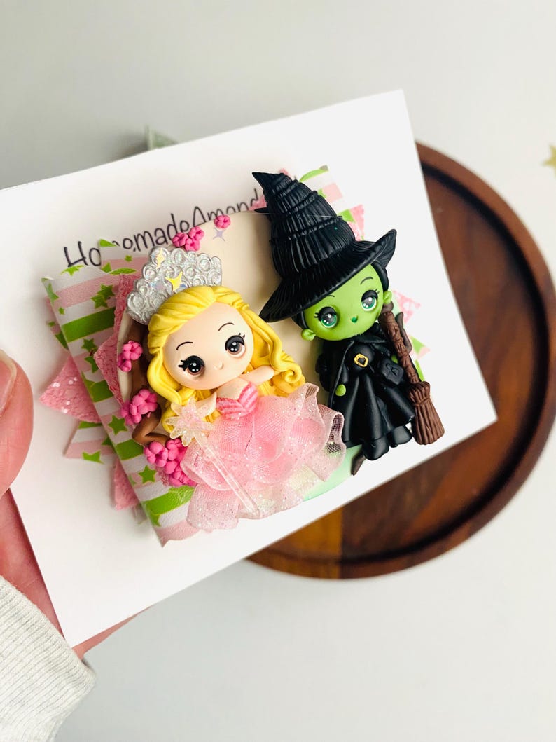 May include: A handmade polymer clay figurine set featuring a pink-dressed princess with a crown and a green witch wearing a black hat and holding a broom. The figurines are on a white card with the text "Homemade Amond".