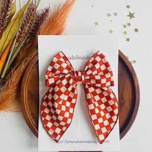 May include: A red and white checkered bow with a white border. The bow is tied with a knot in the center and has a pointed shape at the bottom. The bow is on a white background with the text "https://homemadeamandasaway.etsy.com" at the bottom.