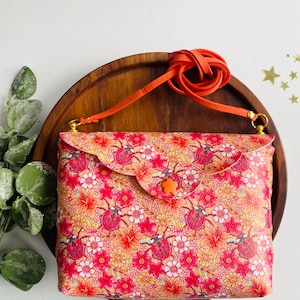 Puede incluir: Un pequeño bolso bandolera de estampado floral naranja y rosa con un patrón de mariquitas. El bolso tiene cierre de solapa y una larga correa naranja.
