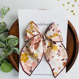 May include: A large floral bow with a cream background and a variety of flowers in shades of yellow, pink, and brown. The bow is on a white card with the text "homemadeamandasway.etsy.com". The bow is trimmed with a burgundy border.