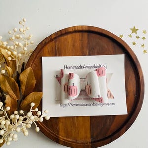 Puede incluir: Un lazo para el pelo blanco y rosa con temática de calabaza sobre una bandeja de madera. El lazo presenta un patrón de calabaza y el texto "HomemadeAmandasWay.etsy.com". Hojas de otoño decorativas y estrellas doradas en el fondo.