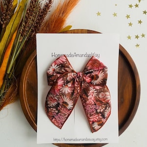 May include: A large, brown, floral patterned hair bow with pink and white flowers. The bow is tied with a loop and is sitting on a white card with the text "HomemadeAmandasWay" and a website address.