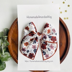 May include: A white bow with a floral print in shades of red, pink, and brown. The bow has a brown border and is displayed on a white card with the text "HomemadeAmandasWay" and a website address.