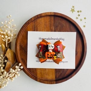May include: A Halloween-themed hair bow featuring Snoopy and a pumpkin. The bow is orange, brown, and gold with a glittery finish. The bow is on a white card with the text "HomemadeAmandasWay" and a website address.