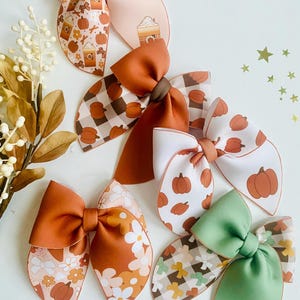 May include: A collection of fall-themed hair bows in various colors and patterns. The bows feature pumpkin, floral, and plaid designs, with shades of orange, brown, and green. Some bows have solid colors, while others showcase printed patterns. The bows are arranged on a white surface.