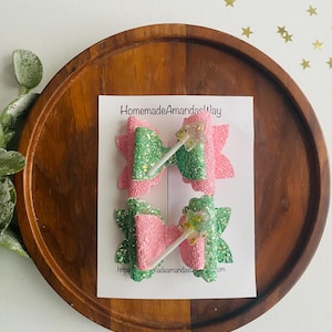 May include: Two pink and green glitter hair bows with a white wand charm. The bows are on a white card with the text "HomemadeAmandasWay" and a website address.