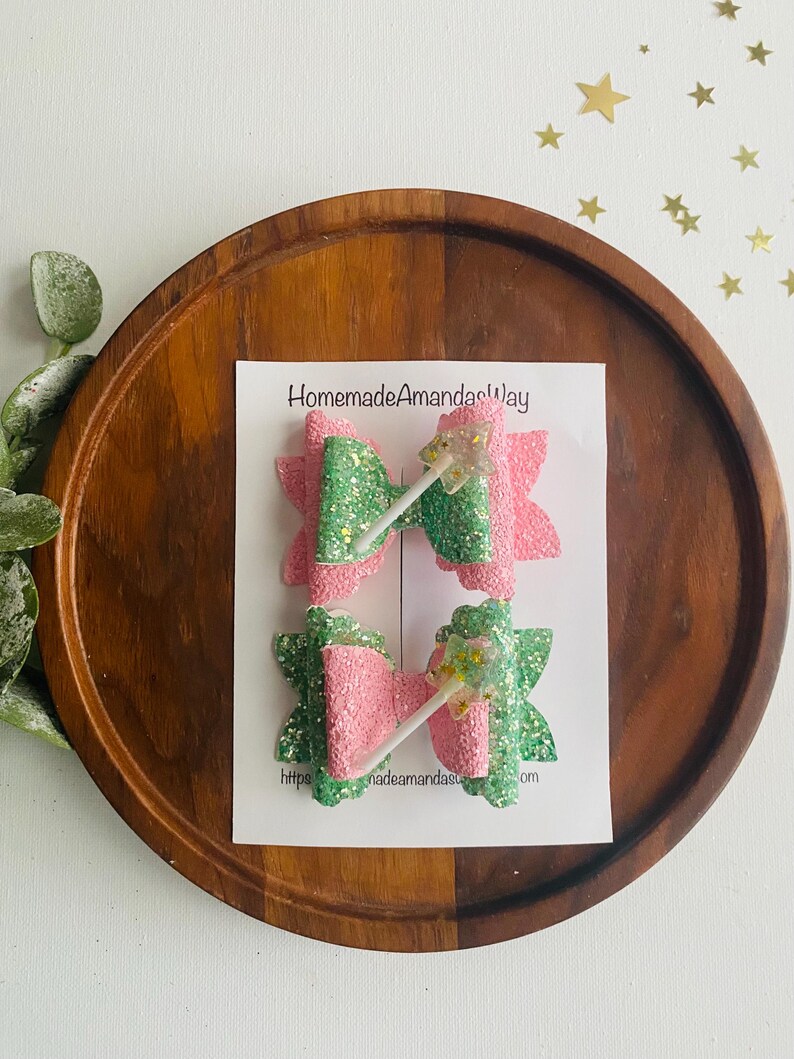 May include: Two pink and green glitter hair bows with a white wand charm. The bows are on a white card with the text "HomemadeAmandasWay" and a website address.