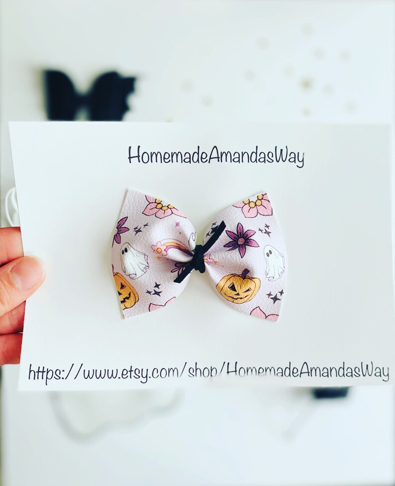 May include: A pink fabric bow with a Halloween theme. The bow features a pattern of pumpkins, ghosts, and stars. The bow is tied with a black ribbon. The text "HomemadeAmandasWay" and "https://www.etsy.com/shop/HomemadeAmandasWay" is printed on the white background.
