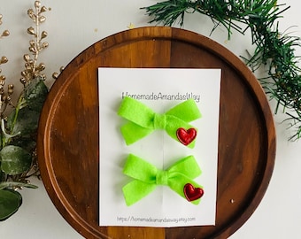 May include: Two lime green felt hair bows with red glitter heart centers. The bows are displayed on a white card with the text "HomemadeAmandasWay" and a website address.