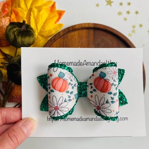 Puede incluir: Un lazo para el cabello hecho a mano con un estampado de calabaza y margarita sobre cuero sintético blanco. El lazo tiene detalles de purpurina verde y está sujeto a una tarjeta blanca con el texto "HomemadeAmanda". La dirección del sitio web también es visible.