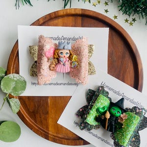 May include: Two handmade hair bows with glitter and a character from a children's book. The first bow is pink with gold glitter and features a princess with a crown and wand. The second bow is green with black glitter and features the Wicked Witch of the West from The Wizard of Oz. Both bows have a website address on the card.