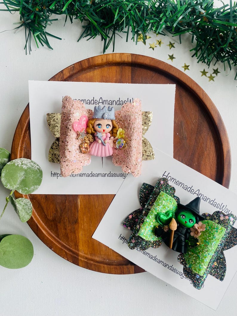 May include: Two handmade hair bows with glitter and a character from a children's book. The first bow is pink with gold glitter and features a princess with a crown and wand. The second bow is green with black glitter and features the Wicked Witch of the West from The Wizard of Oz. Both bows have a website address on the card.