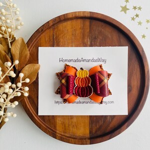 May include: A fall-themed hair bow featuring a layered pumpkin design in shades of orange, burgundy, and gold. The bow is set on a white card with the text "HomemadeAmandasWay" and a website address. The bow is placed on a wooden tray.