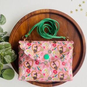May include: A pink crossbody bag with a green strap and a whimsical print featuring witches, hearts, and birds. The bag has a flap closure with a green button.