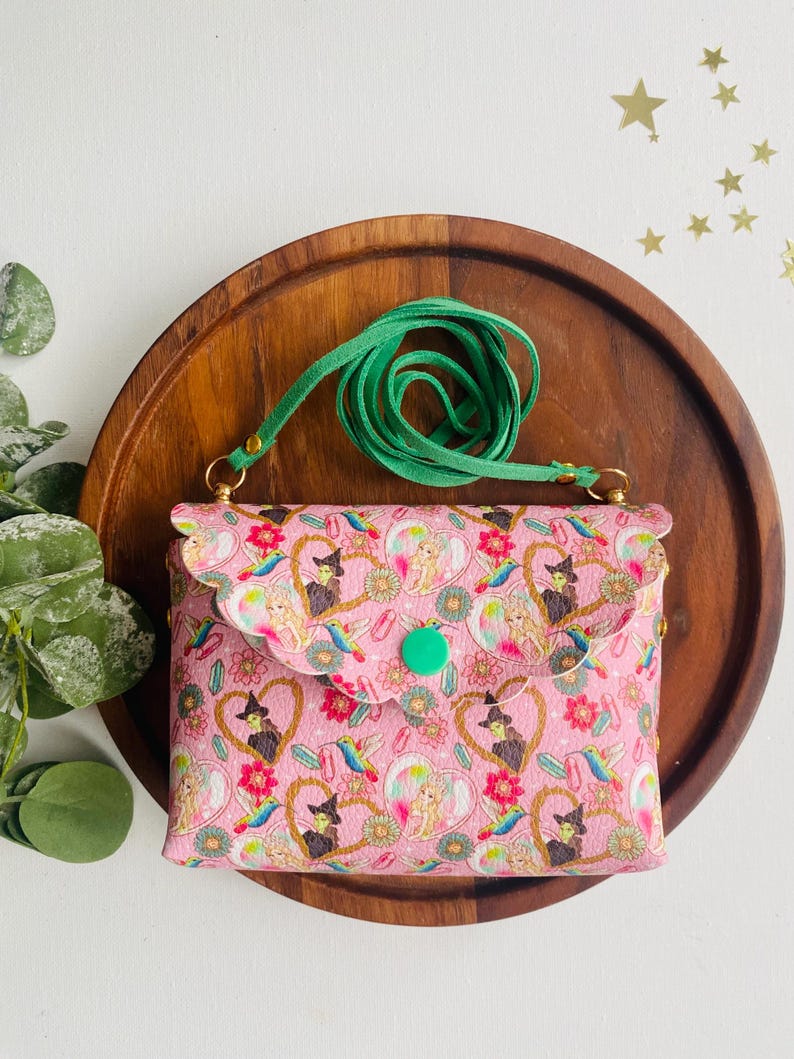 May include: A pink crossbody bag with a green strap and a whimsical print featuring witches, hearts, and birds. The bag has a flap closure with a green button.