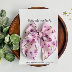 May include: A pink and purple floral hair bow with a light purple trim. The bow is on a white card with the text "HomemadeAmandasWay" and a website address.