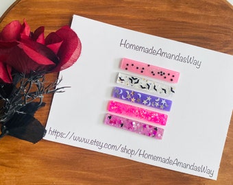 May include: Five colorful hair clips with different Halloween-themed designs, including stars, bats, and moons. The clips are on a white card with the text "HomemadeAmandasWay" and a website address.